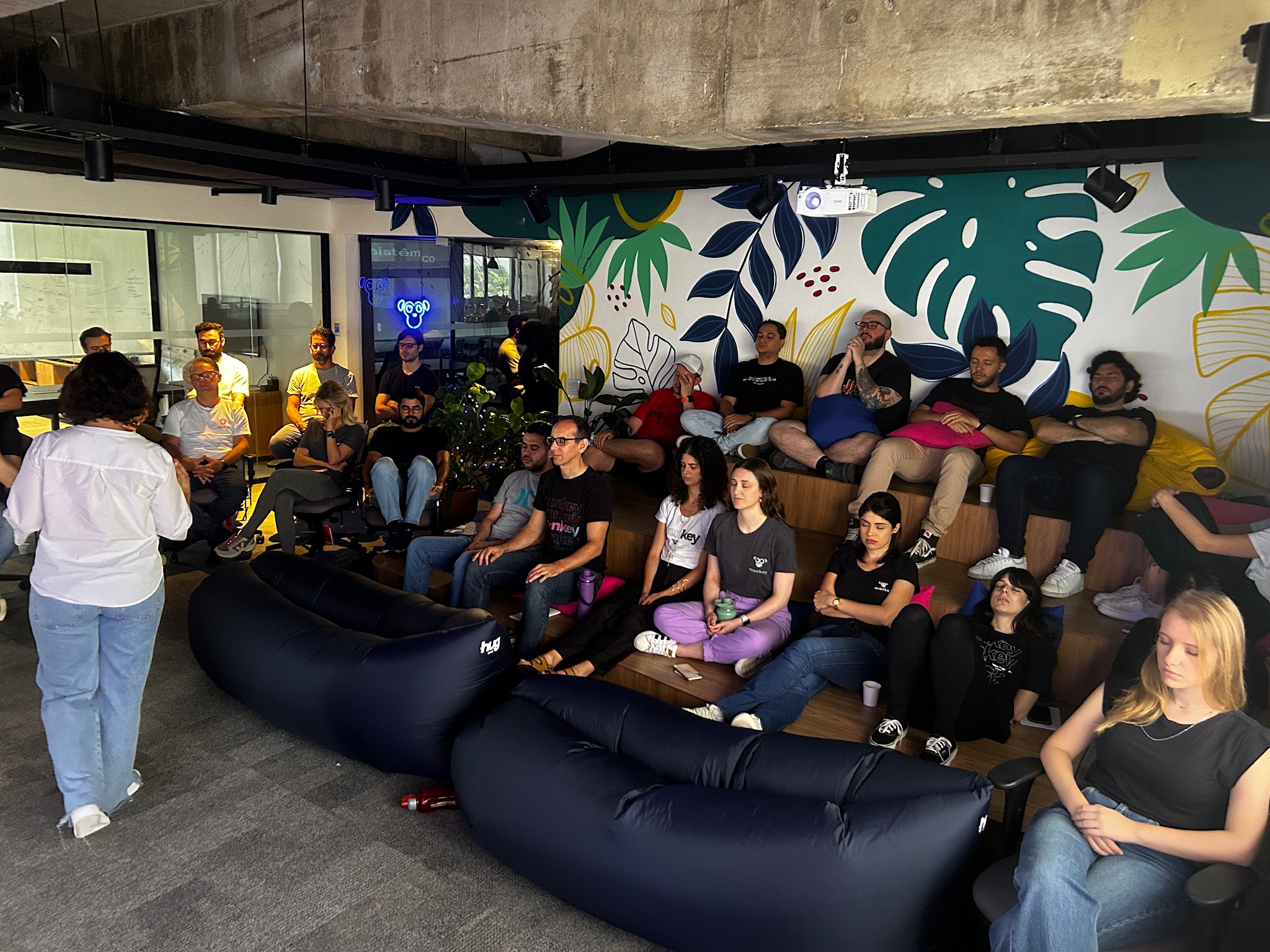 Colaboradores de uma empresa sentados em uma espécie de arquibancada dentro da sede da empresa, com pufes ao redor, todos participando de uma dinâmica de introspecção com os olhos fechados, guiada por uma facilitadora
