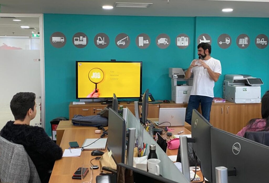 Marcelo Rondon, cofundador da Um Passo Antes, aparece palestrando para um grupo de colaboradores, ao lado de uma TV onde está exposto um PPT com sua fala sobre segurança psicológica