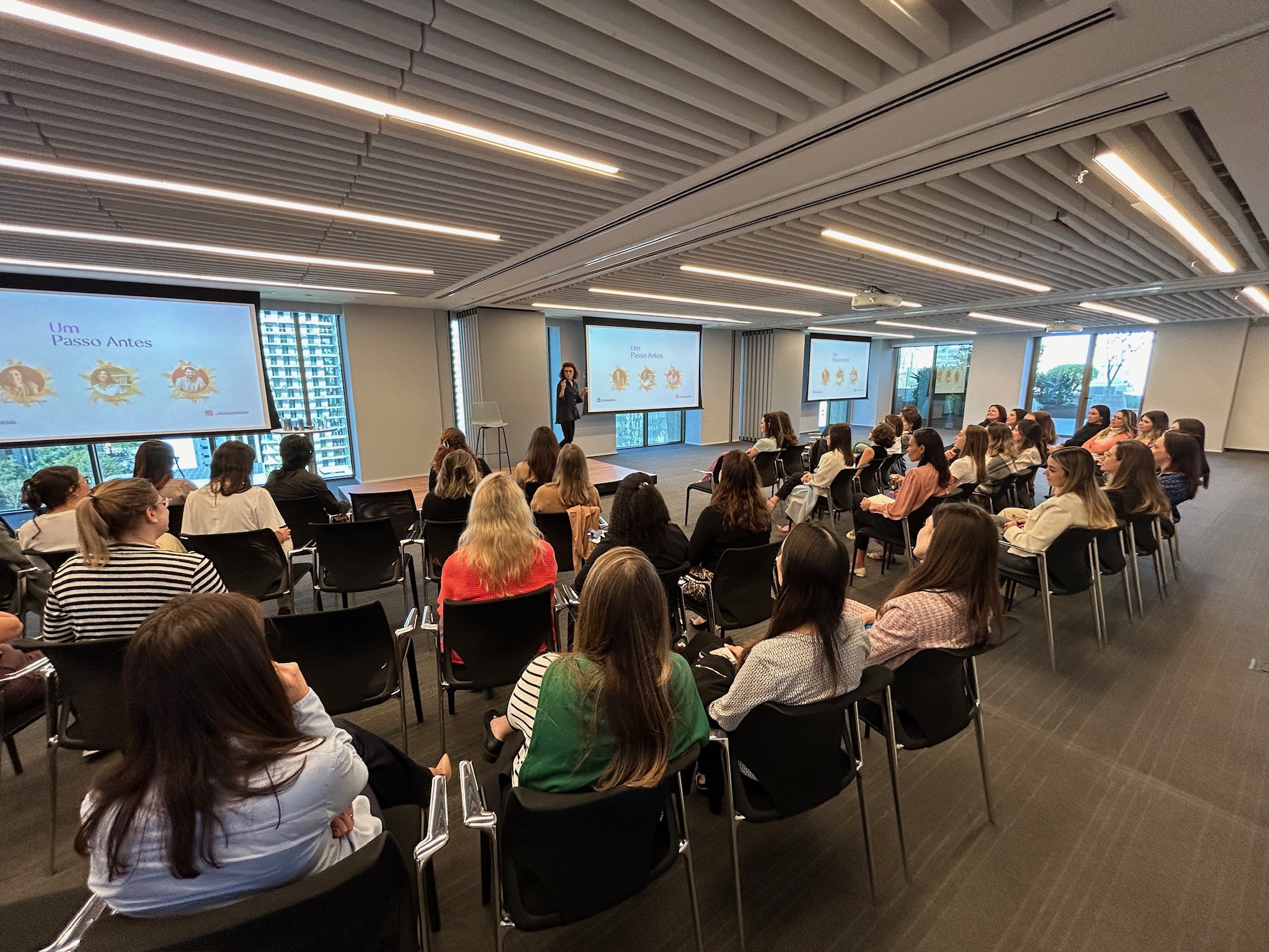 Dezenas de mulheres colaboradoras de uma empresa, vistas de costas, sentadas em cadeiras em forma de meia lua, olhando para o palco onde uma facilitadora conduz sua palestra