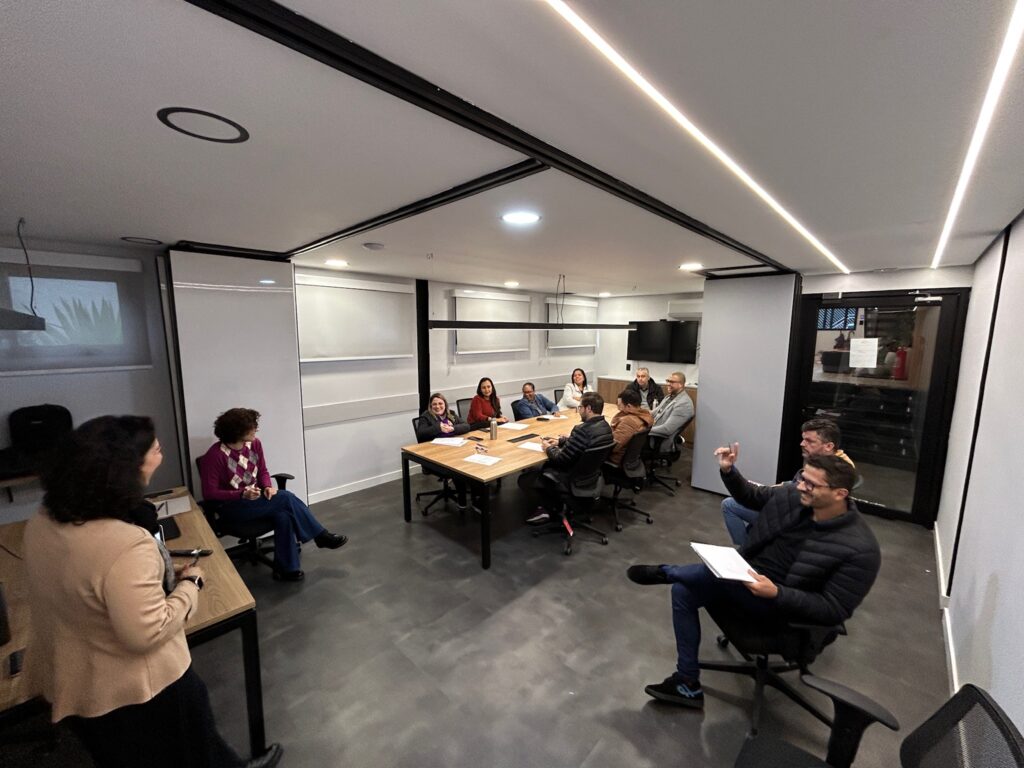 Grupo de colaboradores reunidos em volta de uma mesa de reunião, debatem ideias, guiados por facilitadora em pé