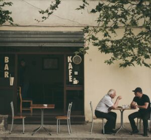 Dois senhores conversam de frente um para o outro, sentados a uma mesa quadrada na calçada, à luz do dia, em frente a uma cafeteria