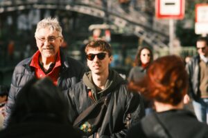 Em uma avenida movimentada, num dia de sol, um homem branco grisalho de óculos caminha ao lado de um jovem branco de óculos escuros. Parecem ser pai e filho